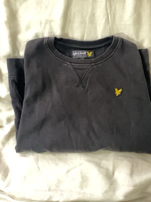 Svart sweatshirt från Lyle & Scott 10/11år - Klassisk svart sweatshirt från Lyle & Scott Junior med rund halsringning och gul logga broderad på bröstet. Tröjan har ribbade muddar vid ärmslut och nederkant, perfekt för en avslappnad och stilren look.