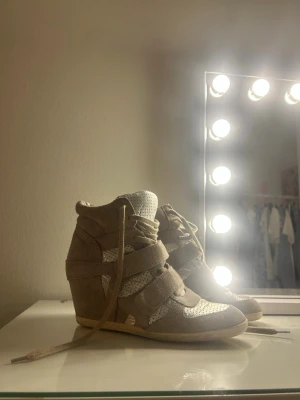 Beige kilklackade sneakers - Snygga beige sneakers med kilklack och detaljer i mocka och mesh. Skorna har snörning och kardborreband för extra stil och passform. Perfekta för dig som vill ha både höjd och streetstyle-vibe i ett par skor. Märket är ASH och liknar stilen av dom populära Isabel marant skorna. Storlek 39 , hör gärna av er vid frågor. Ordinarie pris 2000kr