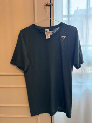 Mörkgrön Gymshark t-shirt - Snygg mörkgrön t-shirt från Gymshark med diskret strukturmönster och liten logga på bröstet. Tillverkad i en mjuk och lätt polyesterblandning som passar perfekt till träning eller en avslappnad stil.