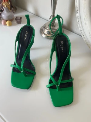 Gröna pumps med remmar från Nasty Gal - Snygga gröna pumps från Nasty Gal med fyrkantig tå och tunna remmar som knyts runt ankeln. Skorna har en modern design med svart innersula och klack. Det saknas ena rem ,