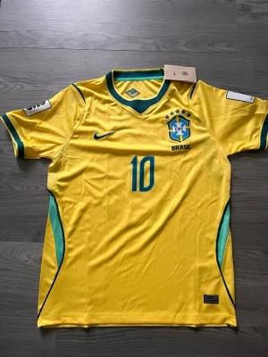 Gul Brasilien fotbollströja nr 10 - Snygg gul Brasilien fotbollströja med nummer 10 och CBF-emblem på bröstet. Tröjan har gröna och blå detaljer vid ärmar och sidor samt rund halsringning. Perfekt för dig som älskar fotboll och vill visa ditt stöd för Brasilien.