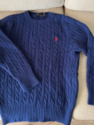 Blå kabelstickad tröja Polo Ralph Lauren - Mörkblå kabelstickad tröja från Polo Ralph Lauren med rund halsringning och röd broderad logga på bröstet. Tröjan har långa ärmar och ribbade muddar vid ärmslut och nederkant. Liten i storlek. 
