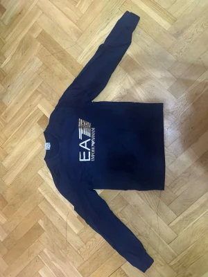 Mörkblå långärmad tröja EA7 - Snygg mörkblå långärmad tröja från Emporio Armani EA7 med tryckt logga i vitt och guld på bröstet. Tröjan har rund halsringning och är tillverkad i ett mjukt material som passar perfekt till en avslappnad stil.