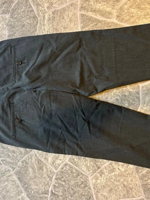 Mörkgrå kostymbyxor 28/32 - Snygga mörkgrå kostymbyxor med raka ben och två bakfickor med knapp. Perfekta för en stilren look. Materialet är mjukt och har en klassisk känsla. Passar dig som vill ha en clean och modern stil.