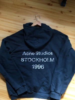 Mörkblå hoodie Acne Studios Stockholm - Navy blue hoodie från Acne Studios med tryck i vitt på ryggen: 'Acne Studios STOCKHOLM 1996'. Klassisk passform med huva och ribbade muddar. Perfekt för dig som gillar stilrena och trendiga plagg.