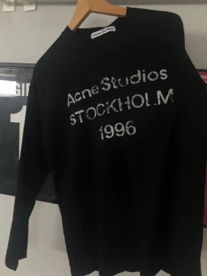 Svart långärmad topp Acne Studios - Cool svart långärmad topp från Acne Studios med vit text 'Acne Studios STOCKHOLM 1996' framtill. Oversized fit och mjukt material, perfekt till jeans eller kjol för en avslappnad look.