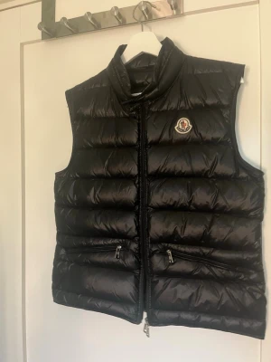 Svart dunväst från Moncler - Snygg svart dunväst från Moncler med glansig finish och klassisk logga på bröstet. Västen har hög krage, två dragkedjeförsedda fickor och quiltad design. Perfekt för lager-på-lager och streetstyle.
