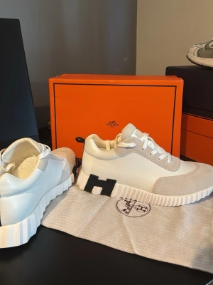 Vita sneakers från H med svarta detaljer - Snygga vita sneakers från H med beige mockadetaljer och svarta inslag på sulan. Skorna har en chunky vit sula med räfflad design och klassisk snörning. Perfekta för dig som gillar exklusiva och trendiga sneakers.