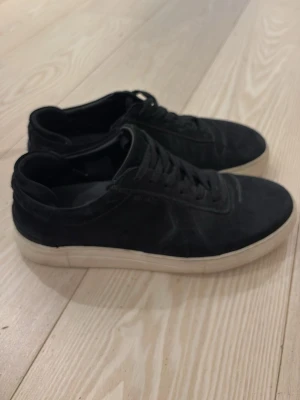 Svarta sneakers från Axel Arigato - Snygga svarta sneakers från Axel Arigato med lågt skaft och vit sula. Skorna har en stilren design med ovandel i mocka och klassisk snörning. Perfekta för dig som gillar minimalistisk och clean stil.