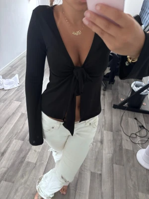 Svart långärmad topp med knyt - Snygg svart långärmad topp med djup v-ringning och knyt framtill. Toppen är croppad och har en figurnära passform som framhäver midjan. Perfekt att styla med jeans eller kjol för en trendig look.