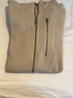 Beige hoodie från Sail Racing - Säljer en beige hoodie från Sail Racing med hel dragkedja framtill och en bröstficka med dragkedja. Hoodien har diskret logga på bröstet och är tillverkad i mjukt material. Perfekt för en avslappnad och sportig stil. Kontakta för intresse!