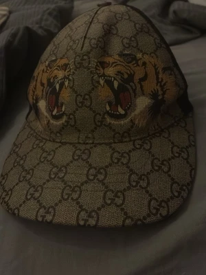 Gucci keps med tigertryck - Säljer en snygg keps från Gucci med klassiskt GG-mönster och två stora tigerhuvuden framtill. Kepsen har böjd skärm, mesh på baksidan och justerbart spänne. Färgerna är beige, svart och detaljer i gult och rött. Perfekt för dig som vill sticka ut.