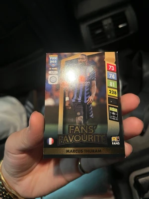 FIFA 365 Marcus Thuram samlarkort - Samlarkort från FIFA 365-serien med Marcus Thuram i Inter-tröja. Kortet har svart och guld design med texten 'Fans' Favourite' och visar spelarens stats. Perfekt för dig som gillar fotboll och att samla på kort.