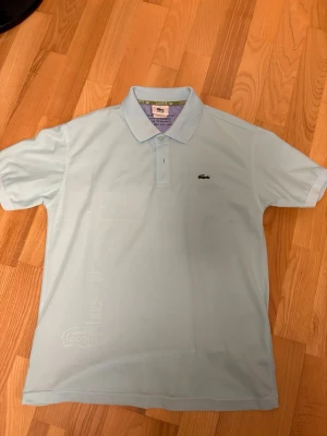 Ljusblå pikétröja från Lacoste - Snygg ljusblå pikétröja från Lacoste med klassisk krage och knappslå framtill. Tröjan har den ikoniska krokodilloggan broderad på bröstet och är tillverkad i ett mjukt bomullsmaterial. Perfekt för en clean och stilren look.