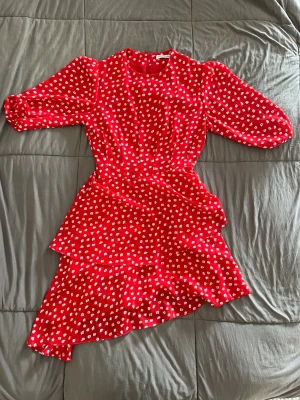 Adoore kläning - Snygg röd kortklänning med vita blommor och omlottdesign. Klänningen har trekvartsärm och volangdetaljer i nederkanten. Perfekt för sommarens alla tillfällen och ger en fräsch och ungdomlig look.
