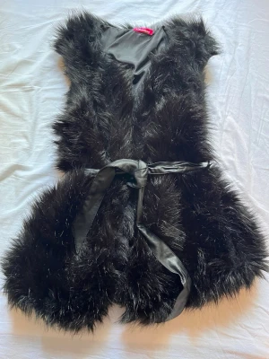 Svart fuskpälsväst från Diabléss - Snygg svart fuskpälsväst från Diabléss med knytband i midjan. Västen har en fluffig look och är helfodrad med slätt tyg på insidan. Perfekt för att lyfta din outfit med en trendig touch.