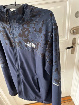 Blå vindjacka från The North Face - Snygg blå vindjacka från The North Face med digitalt camomönster i svart och grått på axlar och huva. Jackan har dragkedja framtill, justerbar huva och logga på bröstet. Perfekt för blåsiga dagar och streetwear-stil.