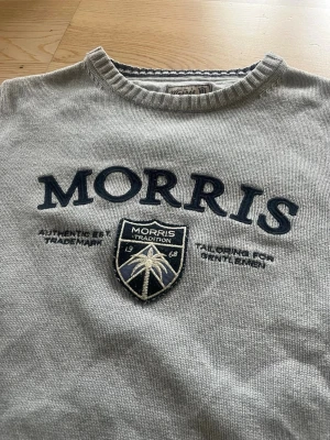 Grå tröja från Morris - Snygg grå tröja från Morris med rund halsringning och broderad logga samt emblem på bröstet. Tröjan har ribbade muddar vid ärmslut och nederkant. Perfekt för en stilren och avslappnad look.