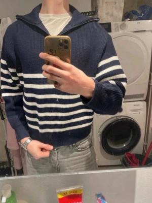 Marinblå stickad tröja med ränder - Säljer en marinblå stickad tröja med vita horisontella ränder och bred krage. Tröjan har avslappnad passform, långa ärmar och ribbade muddar. Perfekt för en casual look och passar till jeans eller chinos. Aldrig använt tröjan! 