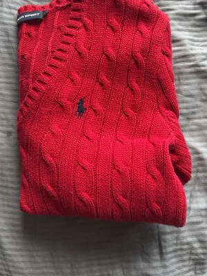Röd kabelstickad tröja Ralph Lauren - Snygg röd kabelstickad tröja från Ralph Lauren med V-ringning och klassisk marinblå logga broderad på bröstet. Tröjan har ribbade muddar och är perfekt för dig som gillar en preppy stil. Materialet ser ut att vara mjukt och bekvämt.