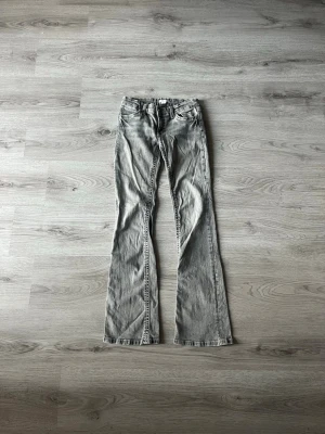 Grå bootcut jeans med slitningar - Snygga grå bootcut jeans med slitna detaljer och klassiska fem fickor. Jeansen har en låg midja och kontrastsömmar längs benen. 