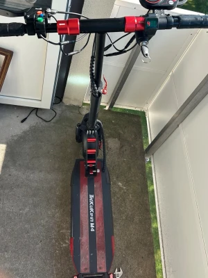 Elscooter Kugoo Kukirin M4 svart/röd - Säljer en elscooter av modellen Kukirin M4 i svart och rött. Scootern har bred fotplatta med röd dekor, LED-display på styret och blåa LED-lampor på sidan. Robust ram i metall och stora hjul för stabil körning. Perfekt för att ta sig fram snabbt i stan.
