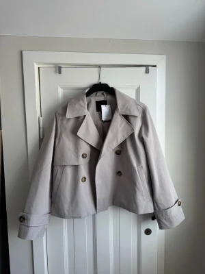 Kort beige trenchcoat med knappar - Ny med prislapp i storlek S. Det innersta knappen har dock lossnat, den medföljer i köpet så att man ska  kunna sy fast den.