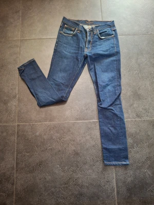 Nudie jeans lean dean - Säljer ett par klassiska blå nudie jeans med kontrastsömmar och fem fickor. Jeansen har normal midja och raka ben, samt snygga detaljer på bakfickorna. Perfekta till en avslappnad stil och passar till det mesta.