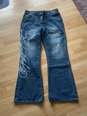 Blå utsvängda jeans med tryck - Säljer ett par blå jeans med utsvängda ben och coolt vitt tryck längs ena benet. Jeansen har slitningar framtill för en trendig look och klassiska fickor både fram och bak. Perfekta för dig som gillar streetwear och vill sticka ut.