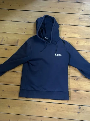 Mörkblå hoodie från A.P.C. - Snygg mörkblå hoodie från A.P.C. med vit logga på bröstet och tryck på ryggen. Klassisk modell med huva och dragsko, samt ribbade muddar vid ärmslut och nederkant. Perfekt för en avslappnad och stilren look. Har två hål som syns på bilderna, men är knappt märkbara i verkligheten. Tveka inte vid frågor.