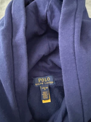 Blå hoodie Polo Bear Ralph Lauren - Säljer en blå hoodie från Ralph Lauren med den ikoniska Polo Bear-trycket på bröstet. Hoodien har huva och ribbade muddar. Tillverkad i mjukt bomullsmaterial och har en avslappnad passform. Perfekt för en chill och stilren look.