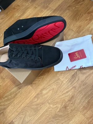 Svarta sneakers med röd sula - Snygga svarta sneakers från Christian Louboutin i mocka med klassisk röd sula och diskret logga broderad på hälen. Skorna har svarta skosnören och en stilren design som passar till det mesta. Kommer med originalpåse och box.