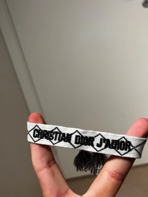 Christian Dior J'Adior tygarmband - Snyggt tygarmband från Christian Dior med texten 'Christian Dior J'Adior' i svart på vit bakgrund. Armbandet har dekorativa tofsar i ändarna och ett grafiskt mönster längs kanterna. Perfekt accessoar för att lyfta din outfit.