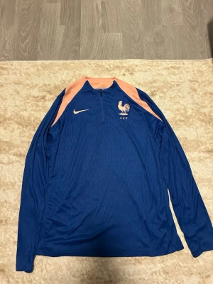Blå Nike träningströja Frankrike - Snygg blå långärmad träningströja från Nike med ljusrosa detaljer på axlarna och halv dragkedja. Frankrikes landslagsemblem med tupp på bröstet. Perfekt för träning eller uppvärmning.