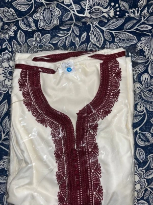 Vit kaftan med vinröda broderier - Säljer en traditionell vit kaftan med detaljerade vinröda broderier längs halsringningen och framtill. Plagget har en lös passform och är tillverkat i ett glansigt tyg som ger en elegant känsla. Perfekt för dig som vill ha något unikt och stilrent.