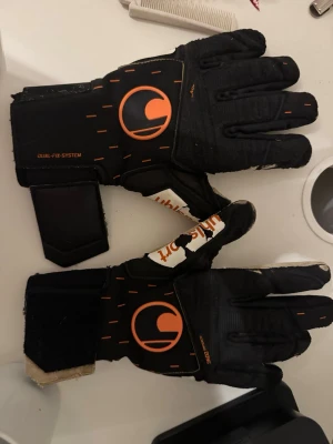 Svarta målvaktshandskar med orange detaljer - Säljer ett par svarta målvaktshandskar med orange logga och detaljer. Handskarna har breda kardborreband vid handleden och greppvänlig insida i beige. Perfekta för dig som spelar fotboll och vill ha bra grepp och stöd.