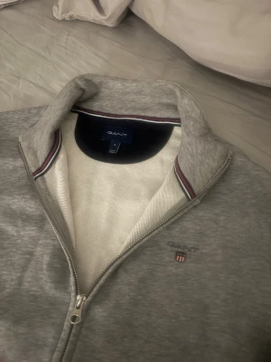 Grå ziptröja från GANT - Snygg grå ziptröja från GANT med hög krage och broderad logga på bröstet. Tröjan har hel dragkedja, ribbade muddar och två stora fickor framtill. Insidan av kragen har en stilig randig detalj. Perfekt för en avslappnad och stilren look.