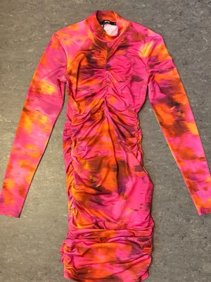 Färgstark långärmad kortklänning - Säljer en supercool kortklänning med lång ärm i starka rosa, orange och svarta toner. Klänningen har rynkade detaljer framtill och på sidorna som ger en snygg form. Materialet känns stretchigt och mjukt, perfekt för dig som vill sticka ut.
