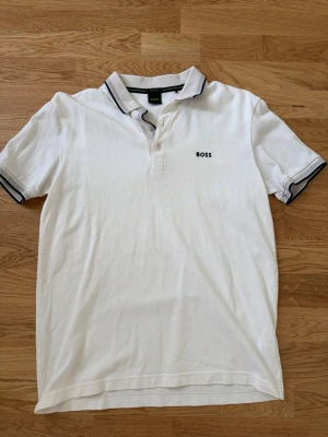 Vit pikétröja från BOSS - Klassisk vit pikétröja från BOSS med svart logga på bröstet. Tröjan har krage med svarta och grå detaljer samt knäppning framtill. Regular fit och kortärmad modell, perfekt till en clean och stilren look.