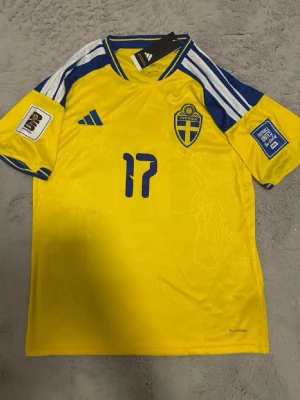 Sverige hemmatröja 2026 - Gul och blå matchtröja från svenska fotbollslandslaget med nummer 17 och namnet Gyökeres på ryggen. Tröjan har blå detaljer, landslagets emblem på bröstet och officiella turneringsmärken på ärmarna. Tillverkad i funktionsmaterial.