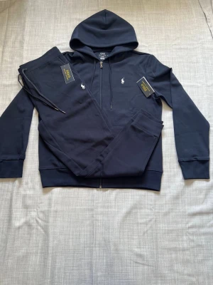 Ralph lauren tracksuit mörkblå strl M  - Helt ny Ralph Lauren tracksuit säljes med hoodie och byxor. Prislapparna är av och Finns en liten defekt mellan benen som ni ser, men inget man lägger märke till.  Väldigt clean och bekvämt set som passar perfekt till vardag och resor. Sjukt populär modell och riktigt bra passform. I storlek L. Snabb frakt och paketpris finns 🔥