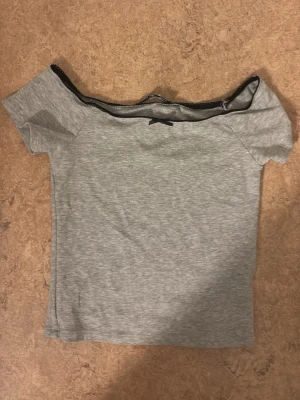 Grå offshoulder topp från FR Sister - Grå offshoulder topp från FR Sister i mjuk bomullsmix. Toppen har korta ärmar, svart spetskant upptill och en liten rosett framtill. Perfekt för en trendig och avslappnad look.