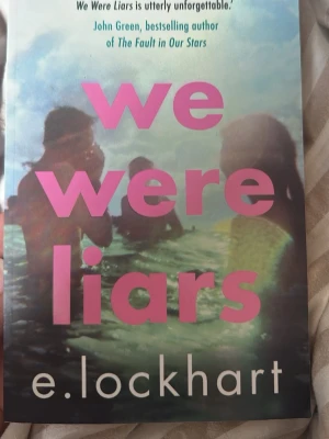 We Were Liars av E. Lockhart - En ungdomsroman på engelska med färgstarkt omslag där rosa text står mot en suddig bild av ungdomar i vatten. Boken är skriven av E. Lockhart och handlar om kärlek, tragedi och hemligheter. Perfekt för dig som gillar spänning och drama.