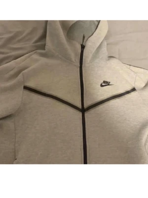 Nike tech dress - Snygg grå hoodie från Nike med hel dragkedja och svart logga på bröstet. Hoodien har svarta detaljer längs dragkedjan och över bröstet, samt en huva. Perfekt för en sportig och avslappnad stil.