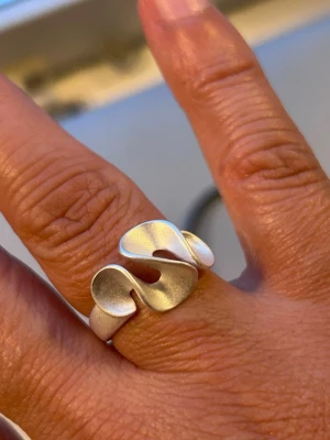 Silvrig ring med vågig design - Unik silvrig ring med en modern, vågig form som ger ett konstnärligt intryck. Ringen är öppen baktill och har en matt finish som gör den stilren och trendig. Perfekt för dig som gillar statement-smycken och vill sticka ut med något annorlunda.