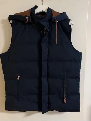Mörkblå dunväst med huva - Snygg mörkblå dunväst med bruna detaljer och avtagbar huva. Västen har dragkedjor på fickorna och knappar framtill. Perfekt för lager på lager och ger en sportig look. Passar till både jeans och hoodie för en chill stil.