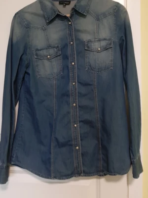 Blå jeansskjorta med tryckknappar - Snygg blå jeansskjorta med långa ärmar och två bröstfickor. Skjortan har klassisk krage och tryckknappar framtill. Perfekt för en avslappnad och cool stil.
