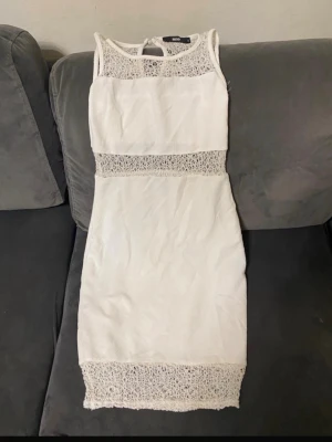 Vit spetsklänning med detaljer - Säljer en vit, ärmlös klänning med spetsdetaljer vid hals, midja och nederkant. Klänningen har en figurnära passform och är knälång. Perfekt för sommarens alla tillfällen och ger en fräsch och elegant look.