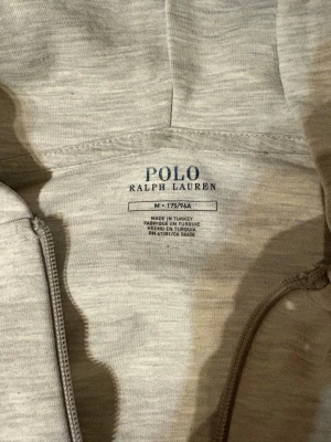 Ljusgrå hoodie från Polo Ralph Lauren - Snygg ljusgrå hoodie från Polo Ralph Lauren med dragkedja framtill, två fickor och klassisk huva med snören. Diskret logga broderad på bröstet. Tillverkad i mjukt material som känns skönt mot huden. Perfekt för en chill och stilren look.