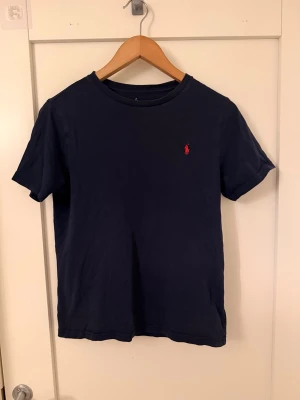 Mörkblå t-shirt med röd logga - En stilren mörkblå t-shirt med rund hals och korta ärmar. På bröstet finns en liten röd broderad logga som ger en snygg detalj. Tillverkad i mjuk bomull för skön känsla och passar perfekt till jeans eller shorts. Trycket inuti med strl är lite bort tvättat men det är en barn L strl och är helt äkta, köpt på kidsbrandstore. Tveka inte på att höra av er vid frågat // Axel H.